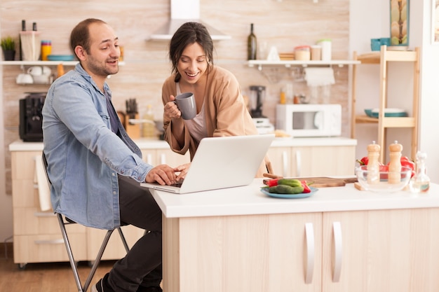 キッチンでラップトップに取り組んでいる起業家と彼の仕事を見ている妻。現代のwifiワイヤレスインターネット技術を使用して自宅で幸せな愛情のある陽気なロマンチックな愛のカップル