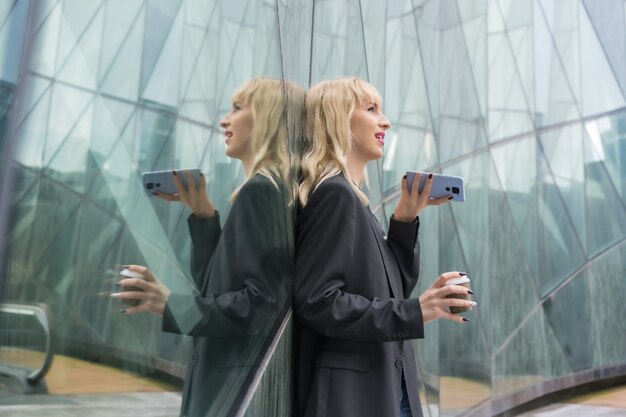 Entrepreneurial girl business park in a modern building reflected in the windows, young blonde in a suit sending a voice message with a take away coffee