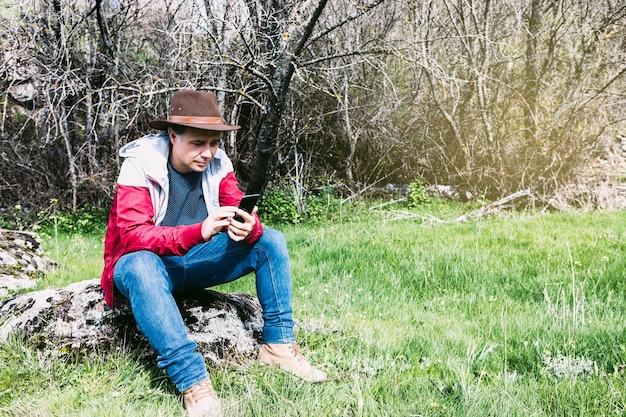 Entrepreneurial selfemployed man wearing hat looking at his mobile while relaxing in the countryside sitting on a stone Concept of work enjoy relax Internet technology and connectivity