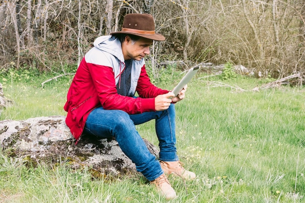 Imprenditore autonomo uomo che indossa un cappello guardando il suo tablet mentre ci si rilassa sul campo seduto su una pietra concetto di lavoro godere di relax tecnologia internet e connettività