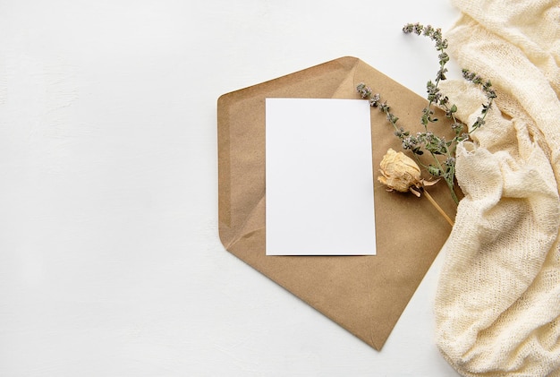 An envelope made of kraft paper a white blank card and dried flowers on a white table Fashionable holiday background top view