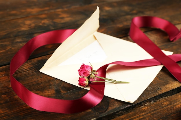 Конверт с любовным письмом, бордовой лентой и сушеной розой на фоне деревенского деревянного стола