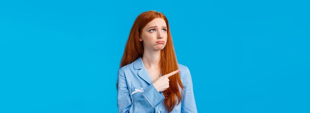 Envy and sad sulking moody redhead teenage girl in nightwear looking and pointing upper left corner