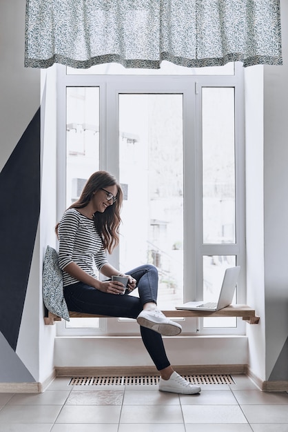 Photo enyoing fresh coffee. beautiful young woman holding a cup and looking at the laptop with smile