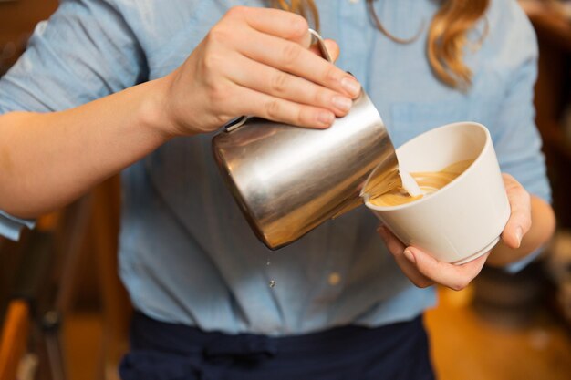 equipment, coffee shop, people and technology concept - close up of woman pouring cream to cup of coffee at cafe bar or restaurant kitchen