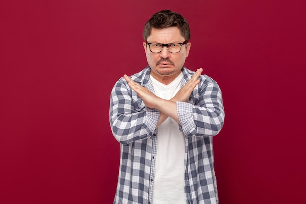 Er is geen manier. Portret van een serieuze, knappe zakenman van middelbare leeftijd in een casual geruit overhemd en een bril die met een X-teken staat en naar de camera kijkt. studio-opname, geïsoleerd op donkerrode achtergrond