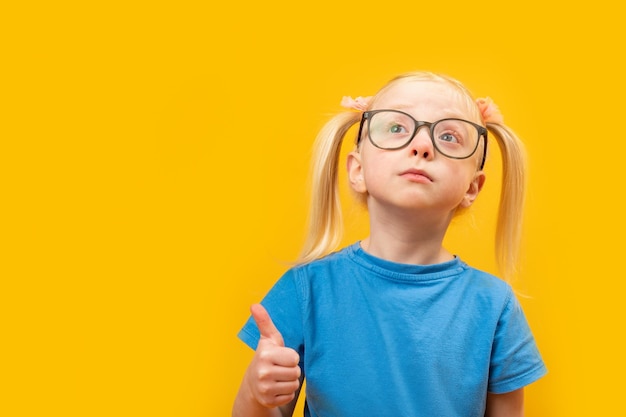 Ernstig basisschoolmeisje met twee paardestaarten draagt een blauwe T-shirt en een bril met de duim omhoog Studioportret van een kind dat een gebaar laat zien