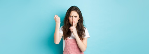 Ernstig en boos brunette meisje schudden vuist en fronsen met afkeuring schelden persoon slecht gedrag vertellen om stil te blijven houden vinger op lippen blauwe achtergrond
