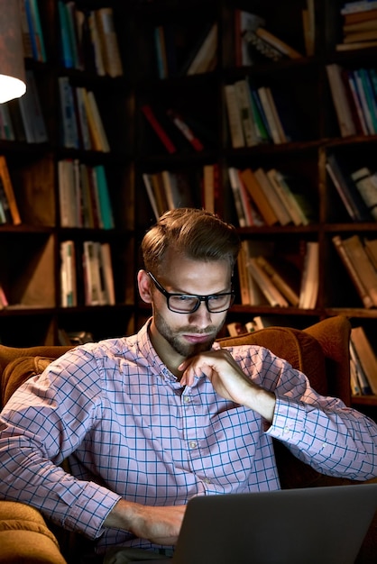 Ernstig gefocust drukke jonge zakenman student die een laptop gebruikt werkt of laat studeert e leert online zit in een stoel kijkt naar een webinar op het kantoor thuis met een boekenkast bij het licht van een nachtlamp