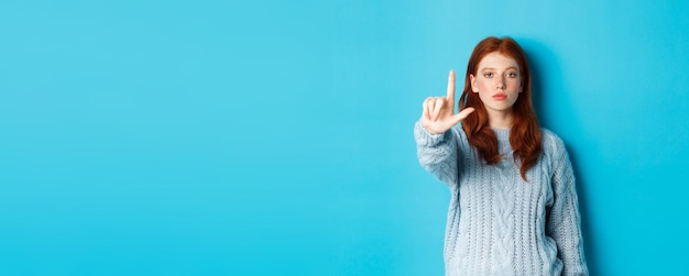 Foto ernstig roodharig meisje in sweater die taboegebaar toont die één vinger uitsteekt en wijsvinger schudt naar dis