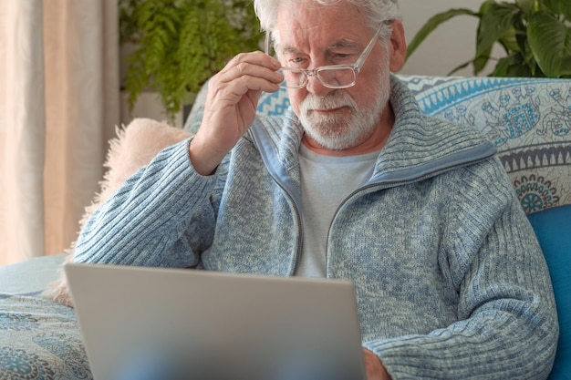 Ernstige 70s senior man zittend op de bank met behulp van laptop business en mensen concept bebaarde blanke oudere man ontspannen thuis werken op afstand