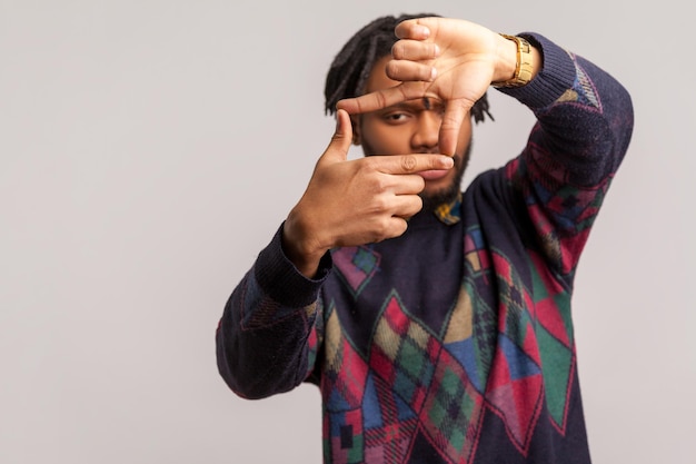 Ernstige afrikaanse man met dreadlocks die door vingers kijkt en cameragebaar maakt, cameramannen die op zoek zijn naar de juiste focus, momenten vastleggen. Indoor studio-opname geïsoleerd op een grijze achtergrond