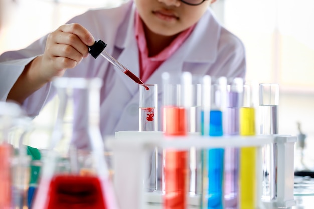 Ernstige Aziatische jongen in laboratoriumjas en bril met druppelaar en buis die kleurrijke reagentia mengt terwijl hij wetenschappelijk onderzoek doet in een modern schoollaboratorium.