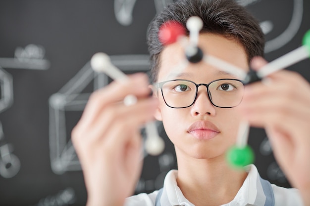 Ernstige Aziatische schooljongen met een bril die in de wetenschapsklas op school staat en naar een plastic molecuulmodel kijkt