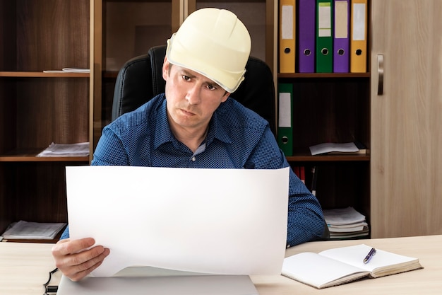 Ernstige baas met helm aan zijn bureau onderzoekt de projectdocumentatie. Bouwer, ingenieur, proctor, voorman.