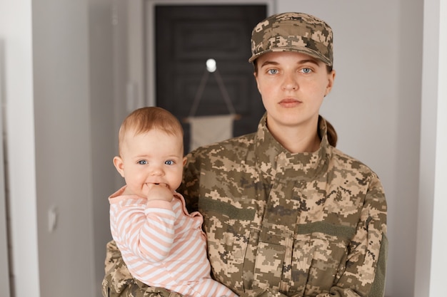 Ernstige bazige militaire moeder die camouflage-uniform en hoed draagt, peuterdochter in de armen houdt en naar camera kijkt met strikte gezichtsuitdrukking.