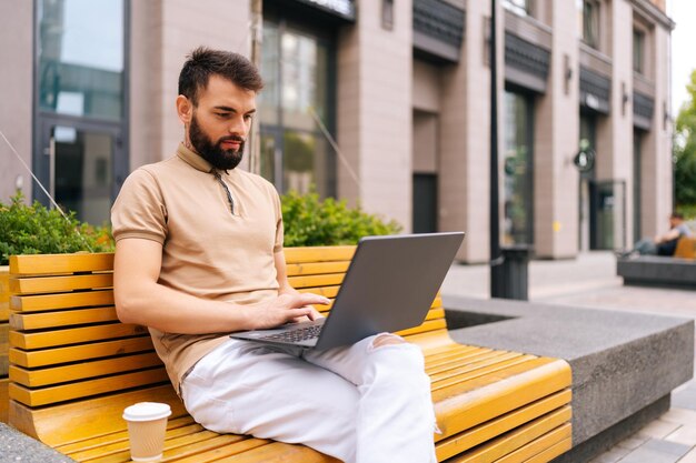 Ernstige bebaarde freelancer man aan het typen met laptopcomputer zittend op een bankje in de straat met afhaalkoffie