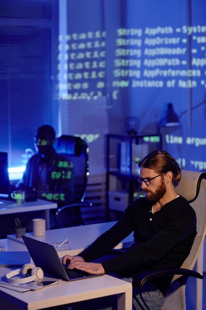 Ernstige, bebaarde jonge zakenman die voor een laptop werkt terwijl hij aan het bureau zit tegen een mannelijke collega in openspace bureau