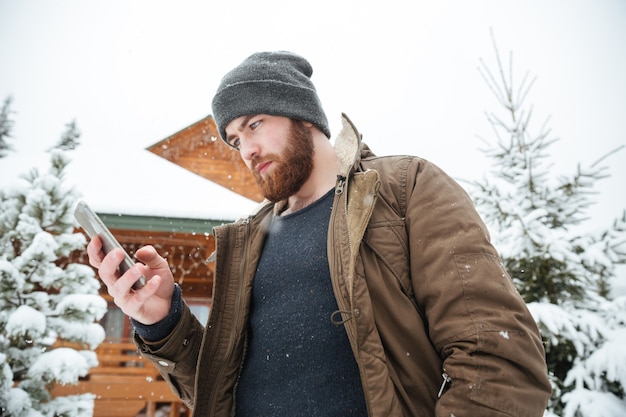 Ernstige bebaarde jongeman die smartphone gebruikt die in de winter buiten staat