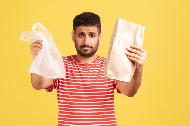 Ernstige bebaarde man in gestreept t-shirt die handen uitsteekt met papier en polyzak, waardoor je de kans krijgt om de juiste keuze te maken en de planeet te redden Indoor studio-opname geïsoleerd op gele achtergrond