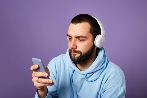 Ernstige bebaarde man luistert naar muziek of podcast in witte koptelefoon met behulp van telefoon