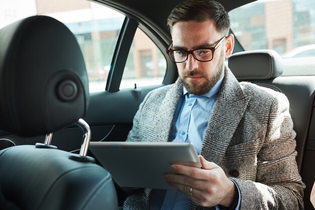 Ernstige bebaarde zakenman in brillen zittend op de achterbank in de auto en met behulp van digitale tablet