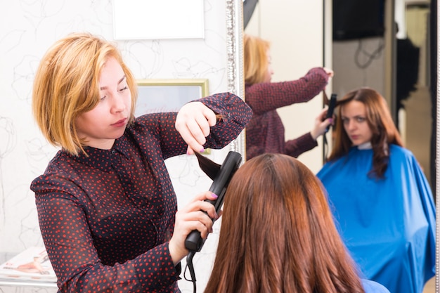 Ernstige blonde stylist met strijkijzer op haar van brunette vrouw in salon met reflectie van cliënt in achtergrondspiegel