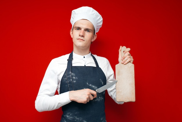 Ernstige chef-kok in uniform houdt een mes en een bord op een rode geïsoleerde achtergrond
