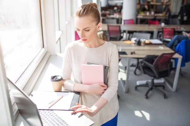 Ernstige en drukke vrouw kijkt naar de laptop en gebruikt het. Ook heeft ze enkele notitieboekjes in haar handen. Vrouw wokring.