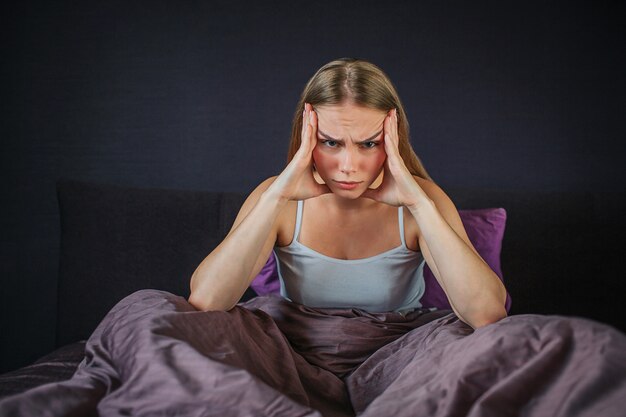 Ernstige en geconcentreerde jonge vrouw zit op bed met gekruiste benen en houdt handen opzij van het hoofd. Ze heeft hoofdpijn. Jonge vrouw lijdt.