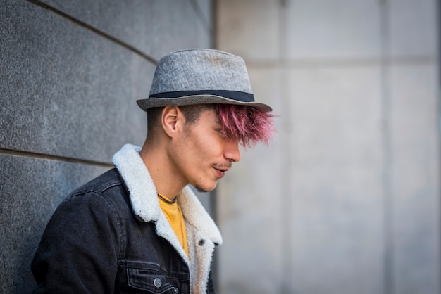 Ernstige en trendy alternatieve stijl tiener man met violet haar en hoed poseren tegen een muur in de stedelijke ruimte. Jongeren in casual spijkerkleding