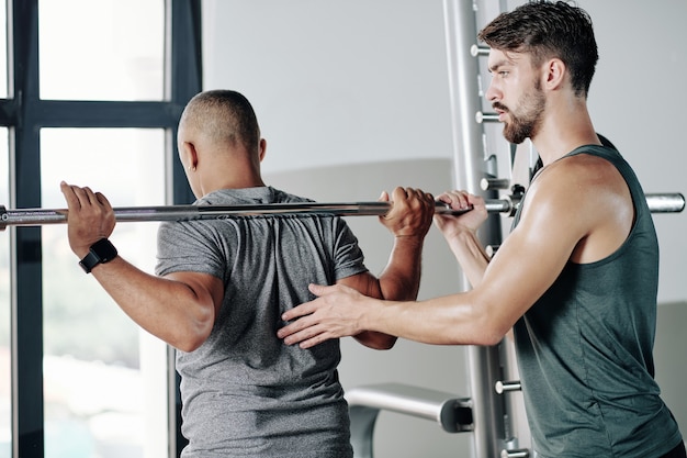 Ernstige fitnesstrainer die cliënt controleert die zich klaar maakt om zware barbell op te heffen