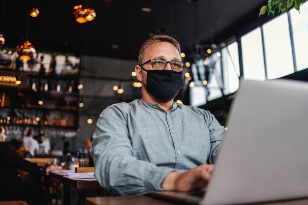 Ernstige freelancer van middelbare leeftijd met bril en beschermend masker op zittend in een café en het gebruik van laptop tijdens corona-uitbraak.