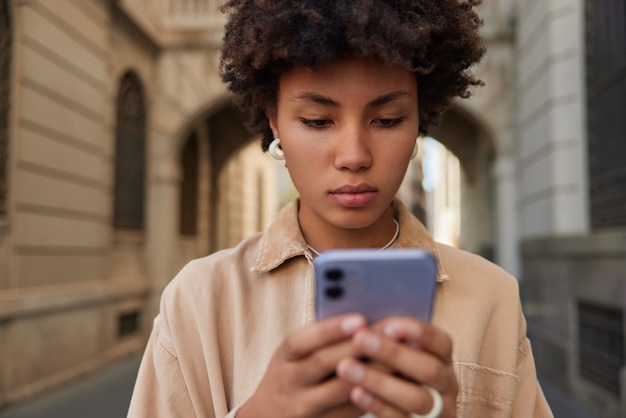 Ernstige gekrulde vrouwelijke blogger maakt nieuwe publicatie in sociale netwerken gebruikt mobiele telefoon voor online surfen geconcentreerd op scherm poses tegen onscherpe achtergrond buiten heeft gateway reizen