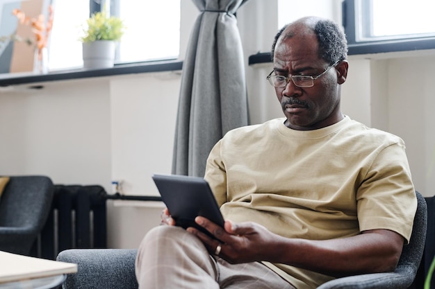 Ernstige gepensioneerde man in brillen en huiskleding die naar het scherm van de tablet kijkt