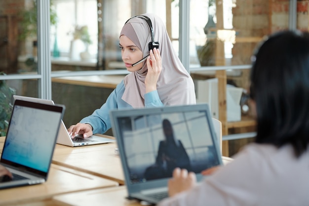 Ernstige jonge Arabische callcenteragent die de microfoon van de headset aanpast terwijl hij reageert op een klant die een laptop op kantoor gebruikt