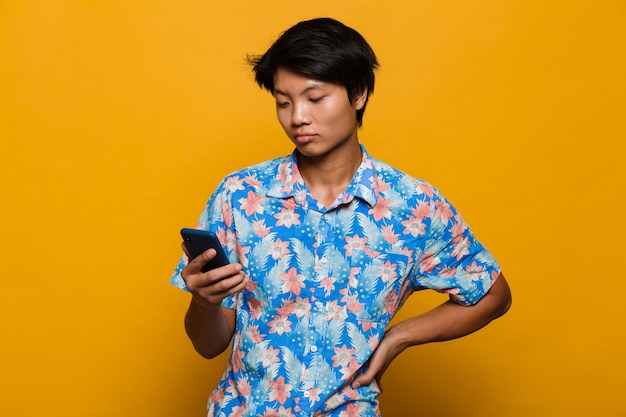 Ernstige jonge Aziatische mens die zich geïsoleerd over gele ruimte bevindt die mobiele telefoon met behulp van.