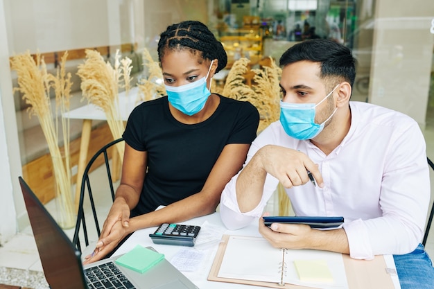 Ernstige jonge bakkerij-eigenaren in medische maskers die financiële rapporten bespreken na de mond van het werk tijdens pandemie