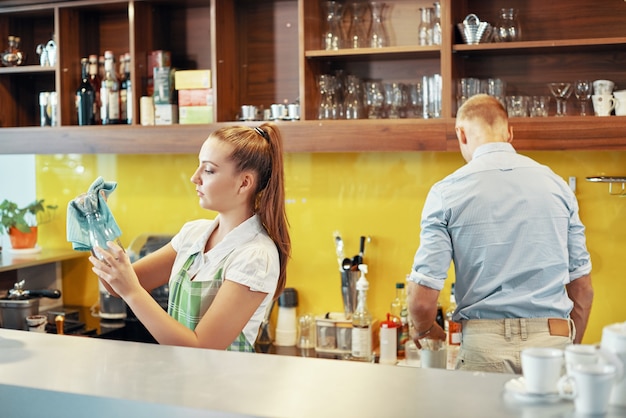 Ernstige jonge barista die glazen potten afveegt met een zachte doek wanneer de manager ingrediënten op de achtergrond bereidt