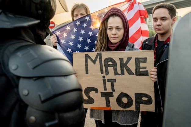 Foto ernstige jonge blanke vrouw met dreadlocks die een spandoek vasthoudt met make stop words die met andere activisten voor de politie in de buitenlucht staat