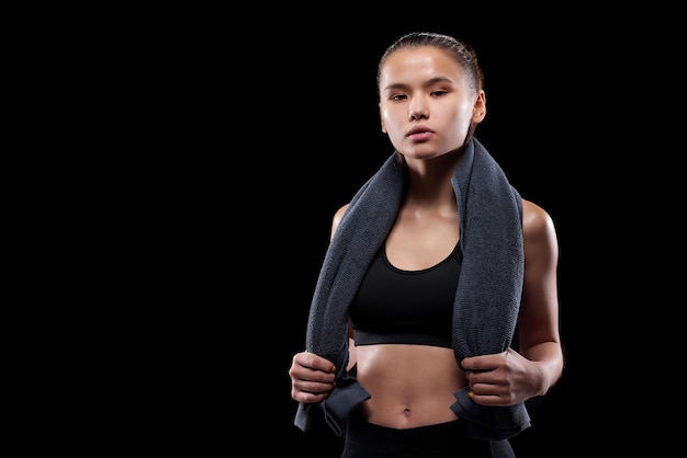 Ernstige jonge geschikte sportvrouw met grijze zachte handdoek op haar hals die zich tegen zwarte muur bevindt