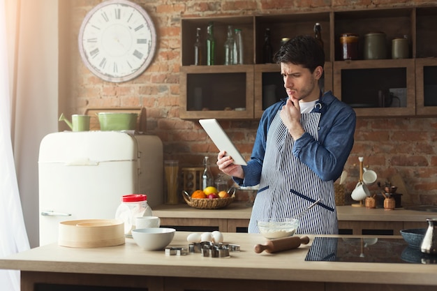 Ernstige jonge man bakt taart in loft keuken thuis, met behulp van digitale tablet met recept. Mockup, kopieer ruimte