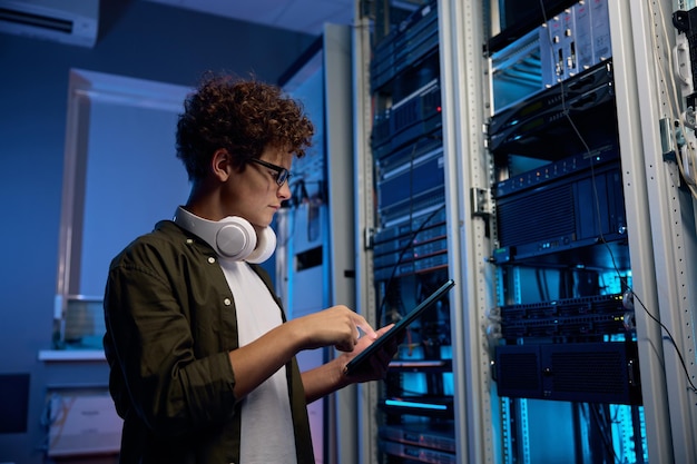 Ernstige jonge man met koptelefoon die in een datacentrum werkt en bij een open server rack cabinet staat en een mobiele tablet gebruikt om het systeem in te stellen