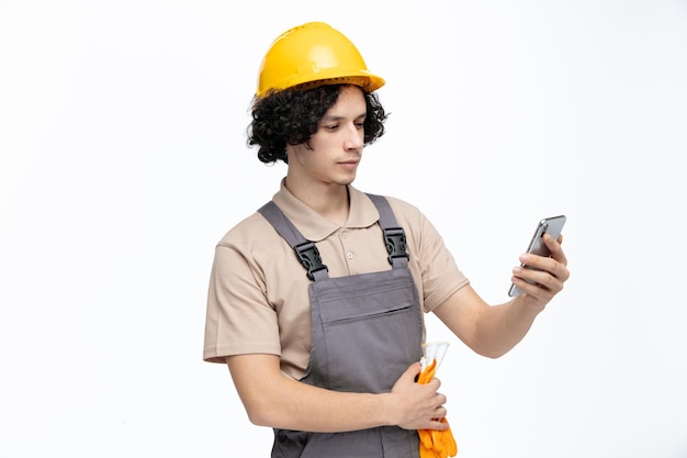 Ernstige jonge mannelijke bouwvakker die uniforme veiligheidshandschoenen en mobiele telefoon draagt die naar mobiele telefoon kijkt die op witte achtergrond wordt geïsoleerd