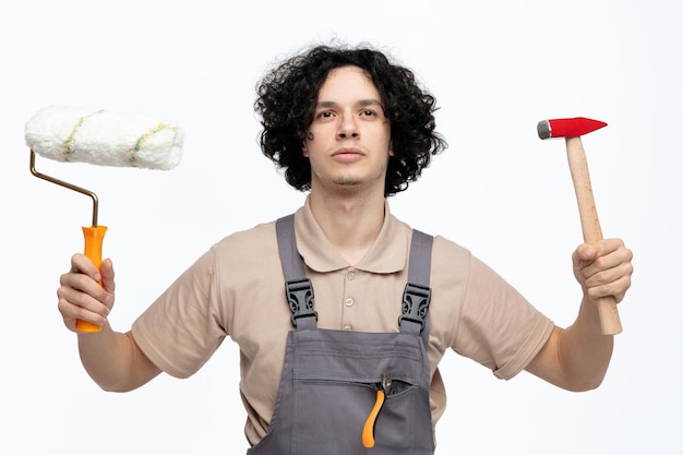 Ernstige jonge mannelijke bouwvakker dragen uniform opzoeken met hamer en verfroller in zijn handen met een tang in zak geïsoleerd op een witte achtergrond