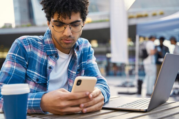 Ernstige jonge Midden-Oosterse man met een bril die een smartphone vasthoudt, met behulp van een laptopcomputer