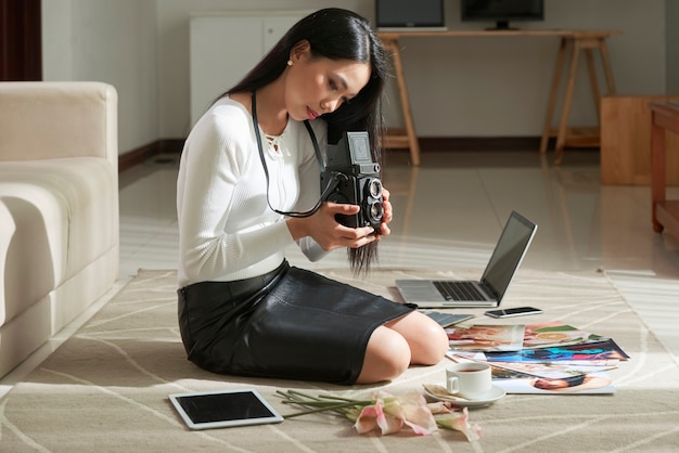 Ernstige jonge Vietnamese fotograaf met lang haar zittend op tapijt met foto's en apparaten en afbeeldingen kiezen op vintage camera