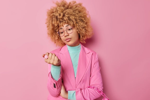 Ernstige jonge vrouw met krullend haar kijkt aandachtig naar haar nagels voelt zich verveeld terwijl ze wacht op iets gekleed in elegante kleding geïsoleerd over roze achtergrond denkt aan het maken van nieuwe manicure