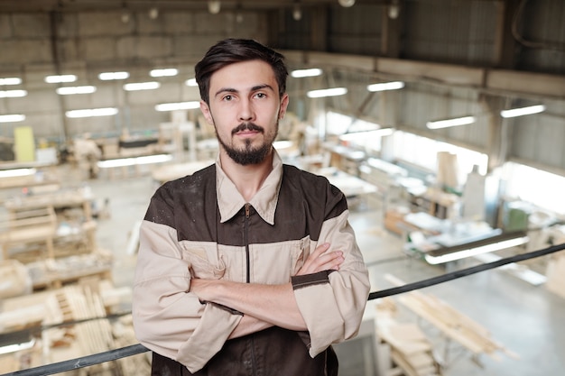 Ernstige jonge werknemer in uniform met zijn armen gekruist door borst staande tegen interieur van grote werkplaats van fabriek