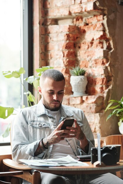 Ernstige jongeman in spijkerjasje zittend aan tafel in loft café en het gebruik van smartphone tijdens het sms'en van sms